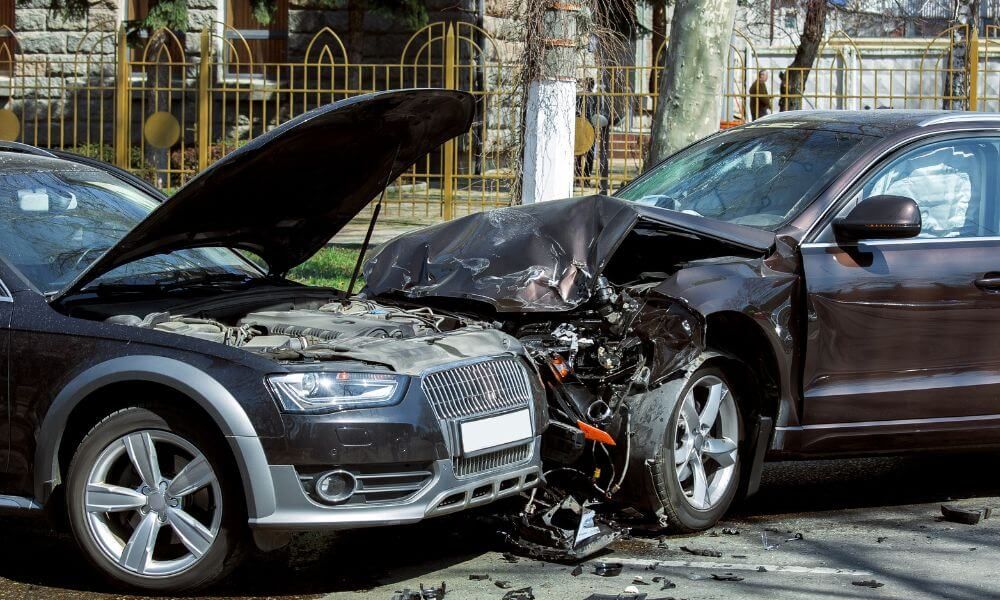 Two cars are involved in a collision; one has its hood raised, exposing the engine, while the other has significant front-end damage. The scene is set on a paved street with a fence in the background.
