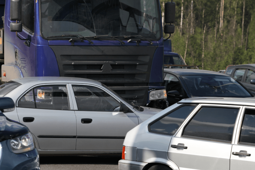 A large blue truck is stationary amidst several smaller cars, including a silver sedan and a gray hatchback, all condensed together in a busy, possibly congested area surrounded by greenery.