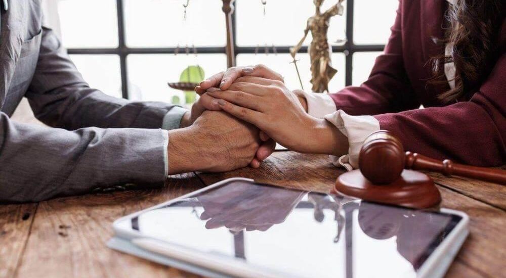 Two professionals clasp hands in agreement across a wooden desk, with a gavel, scales, and a tablet nearby in a legal office setting