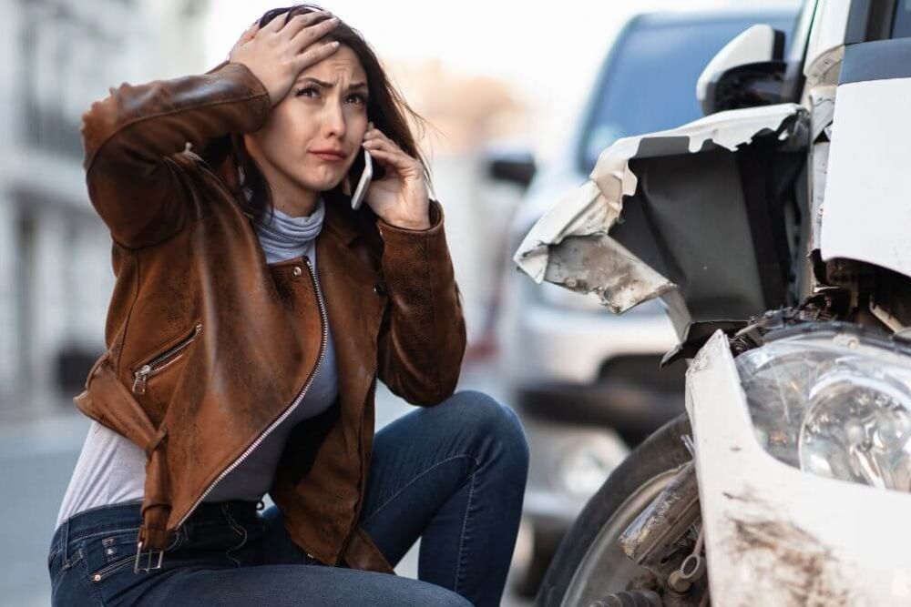 A woman in a brown jacket crouches beside a damaged car, looking distressed while speaking on her phone. She is in a street with other vehicles in the background.