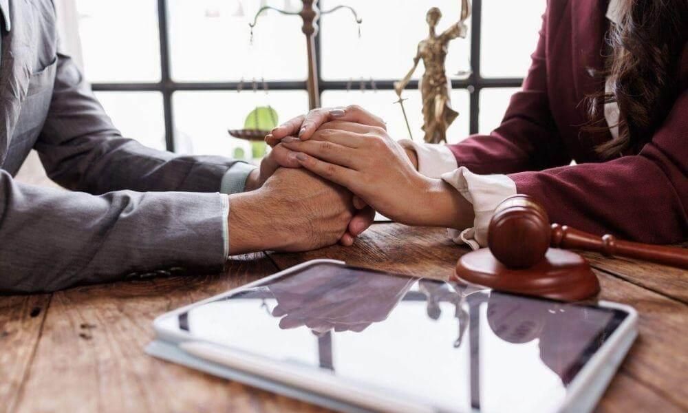 Two professionals clasp hands in agreement across a wooden desk, with a gavel, scales, and a tablet nearby in a legal office setting