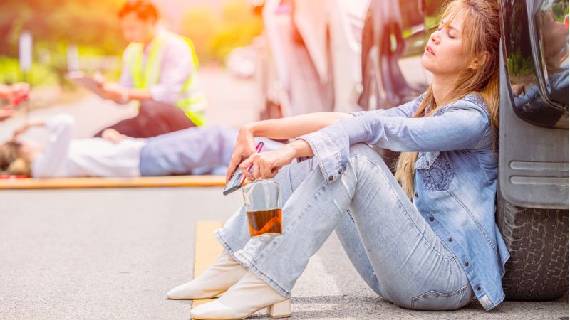 Woman sits on the road beside a vehicle, resting with eyes closed, holding a glass of orange drink while responders work in the background.