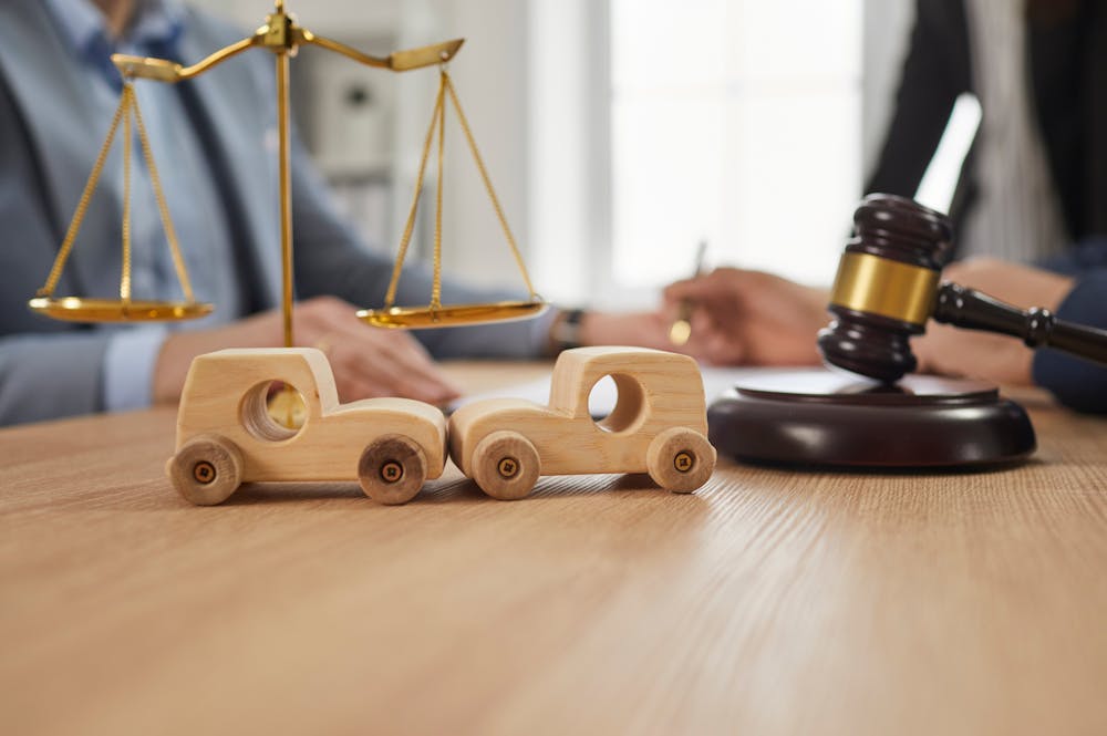 A wooden scales of justice stands above two wooden toy cars on a table. In the background, hands are seen signing documents, indicating a legal context. A gavel rests nearby.