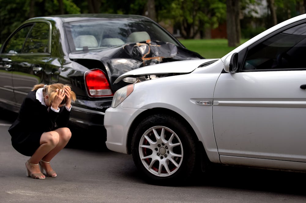 A woman in formal attire crouches on the ground, visibly distressed, next to two damaged cars parked on a road surrounded by trees, indicating a recent car accident.