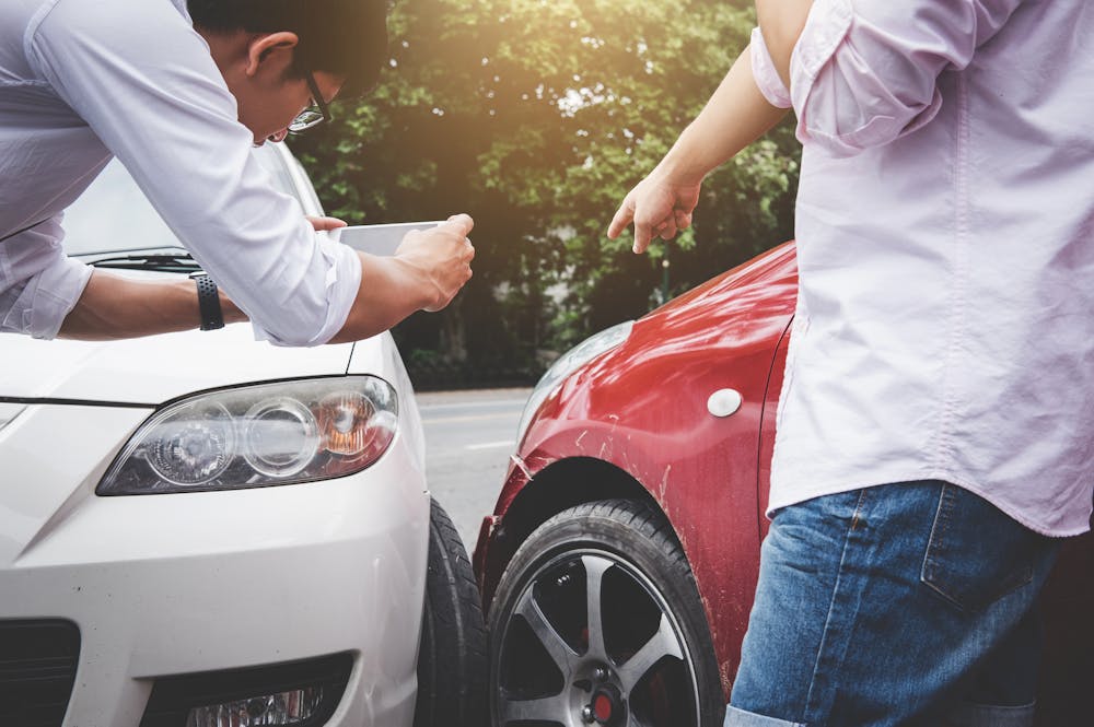 A person in a white shirt photographs a minor car accident between a white and a red vehicle, while another person gestures, discussing the situation amidst a tree-lined background.